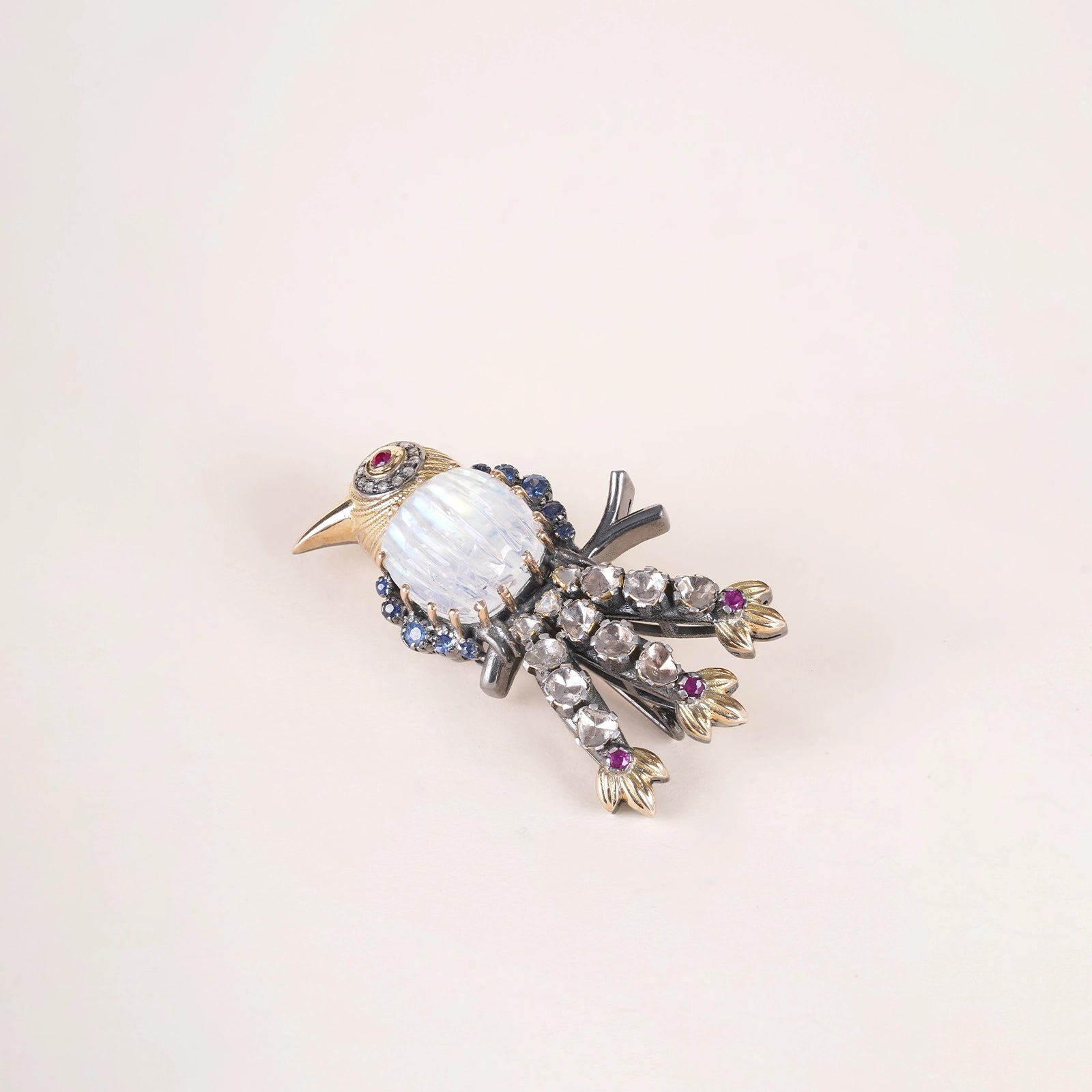 Lovebird Sky Brooch featuring a carved moonstone bird with blue sapphire detailing, ruby eye, uncut diamond tail feathers, and pearl accented chain in gold.